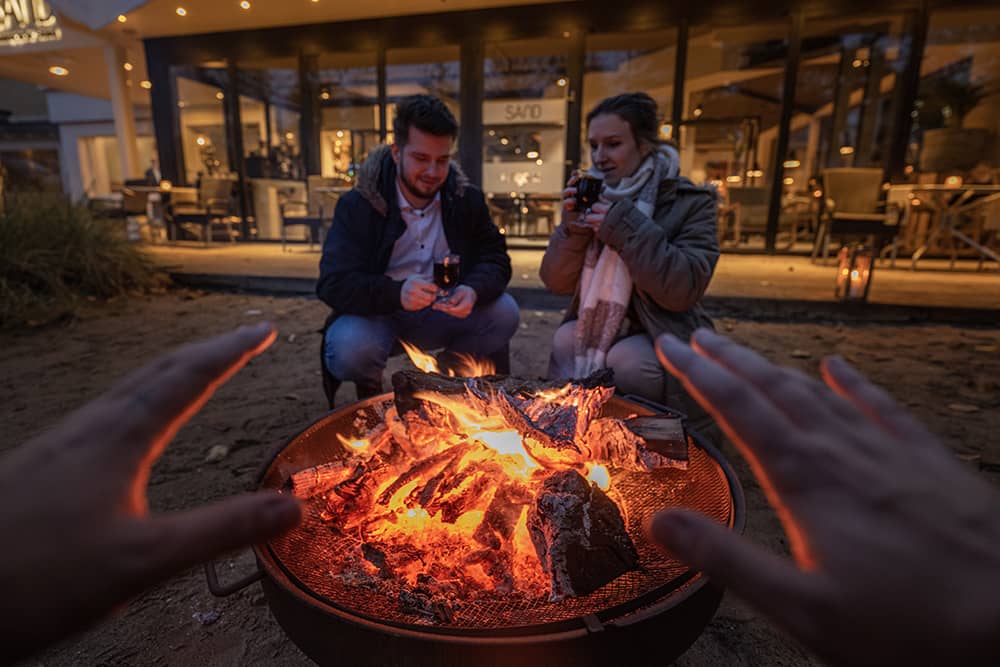 Ein lodernder Feuerkorb auf einer Sand Terrasse an dem ein junges Paar Glühwein trinkt.