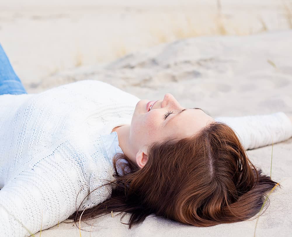 A young woman lying in the sand with her arms outstretched, looking up at the sky.