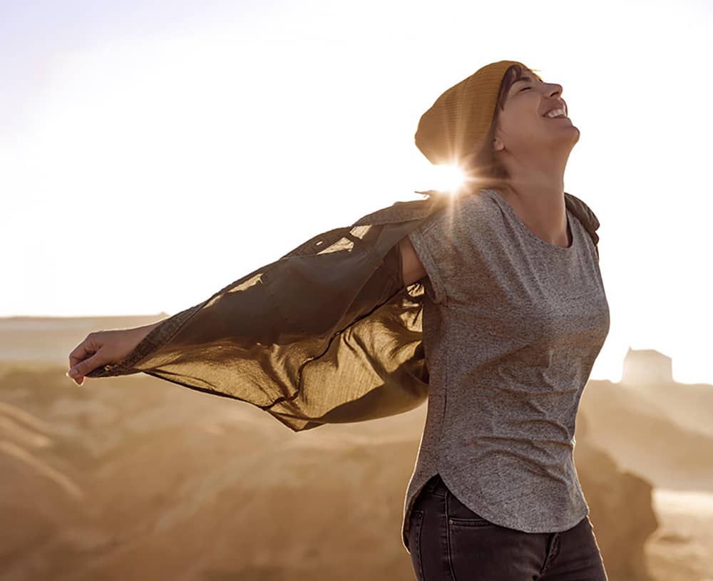 A radiantly laughing young woman with outstretched arms in front of a blazing sun and sandy backdrop.